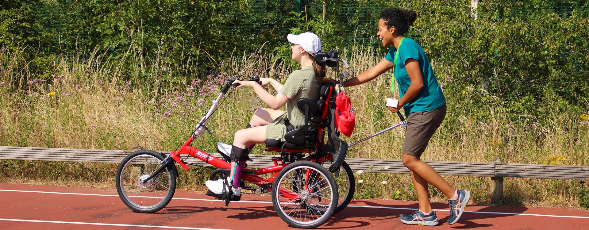 A physiotherapist pushes Treloar's student in an adapted tricycle along a track surrounded by greenery. It is a hot day, both women are wearing shorts and T-shits.