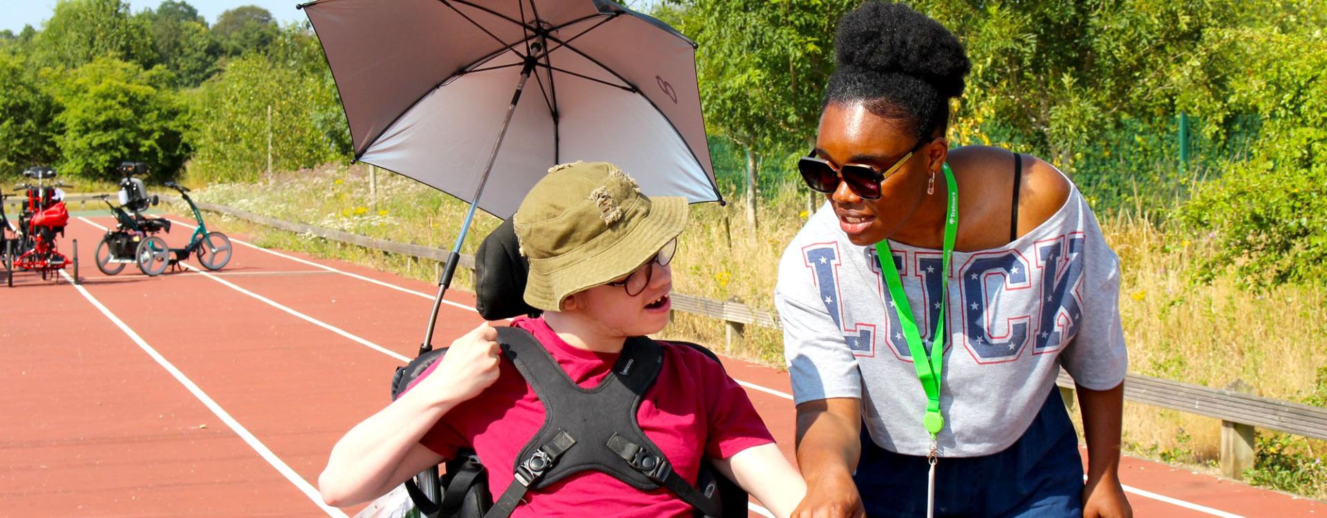Treloar College student with an umbrella attached to his wheelchair on a sports track chatting with his assistant who is smiling and wearing sunglasses. It is a sunny, warm day.