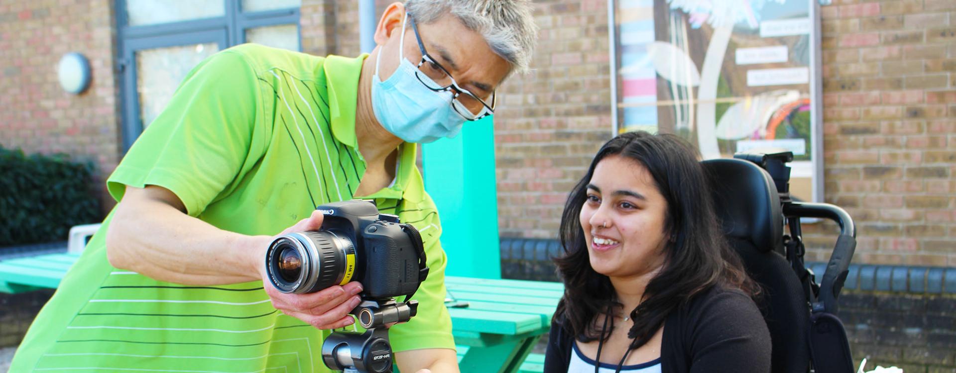 Treloar's student support assistant wearing a mask adjusts a DSLR camera on a tripod while Treloar College student watches him and smiles. they are outdoors, there is a brick wall behind them.
