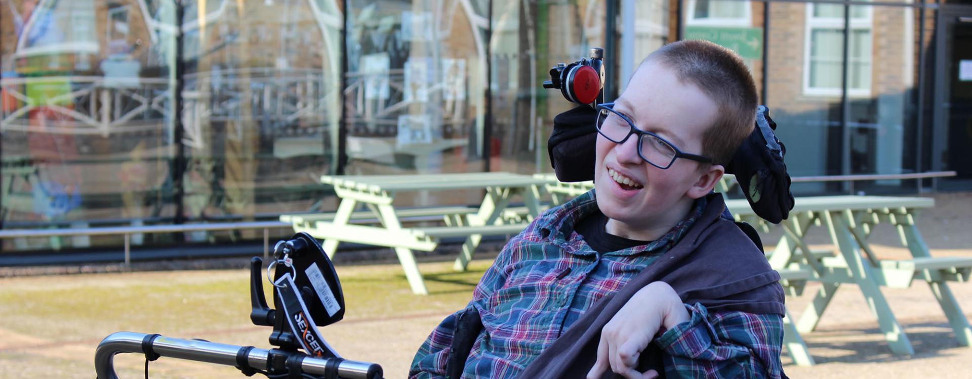 Treloar College student smiling outdoors near picnic tables and a glass building. He is wearing glasses and looks happy.