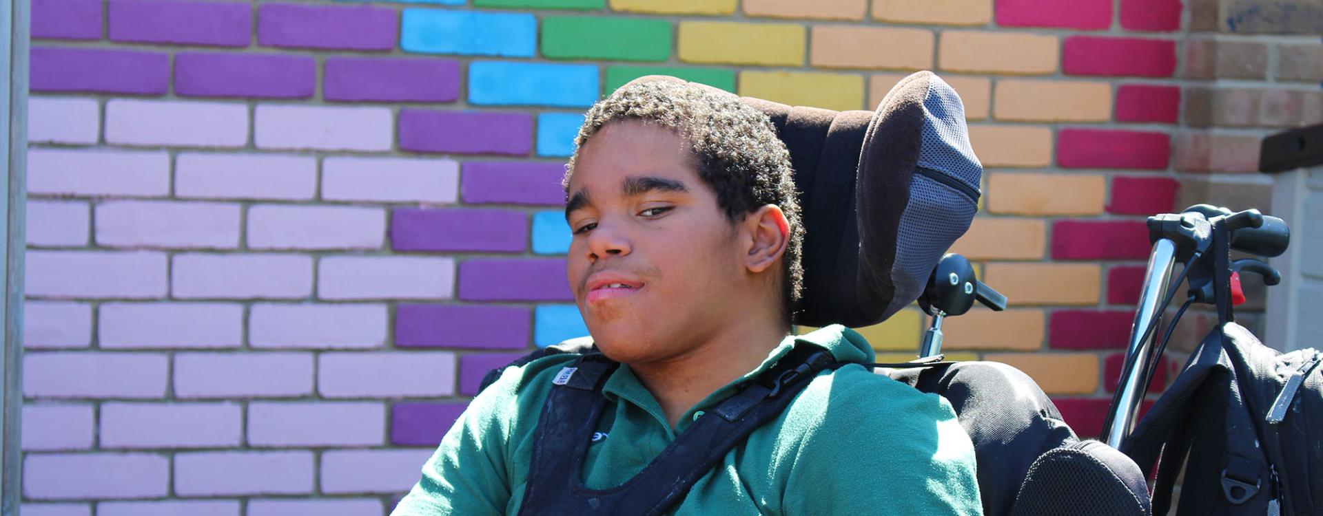 Treloar School student wearing a green T-shirt uniform and poses in front of a colourful brick wall painted in purple, blue, yellow and red hues.