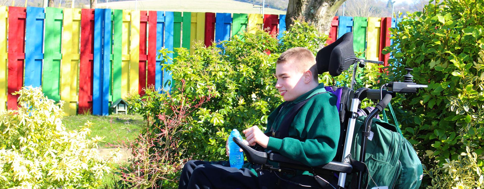 Treloar School student enjoying a sunny day in a garden with colourful wooden fence in the background. He is practising driving his wheelchair outdoors.