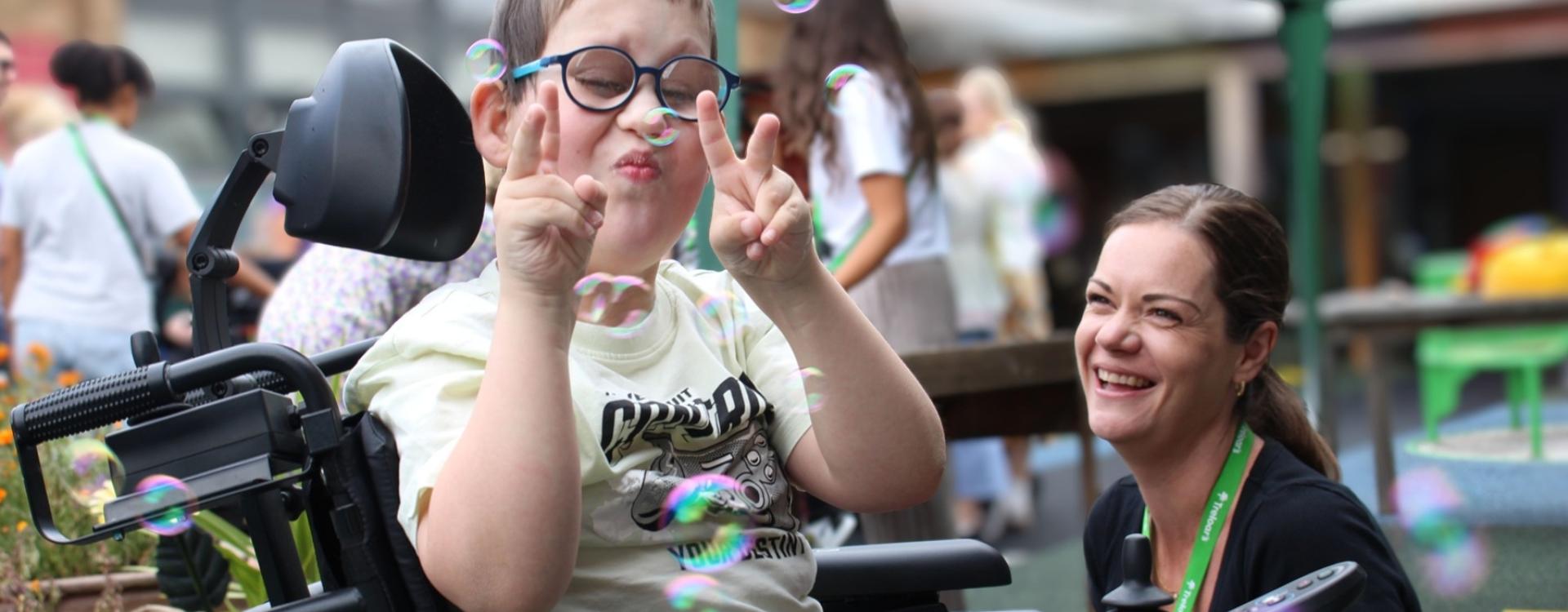 Treloar's primary student making peace signs while surrounded by floating bubbles at an outdoor event with his assistant next to him, smiling.