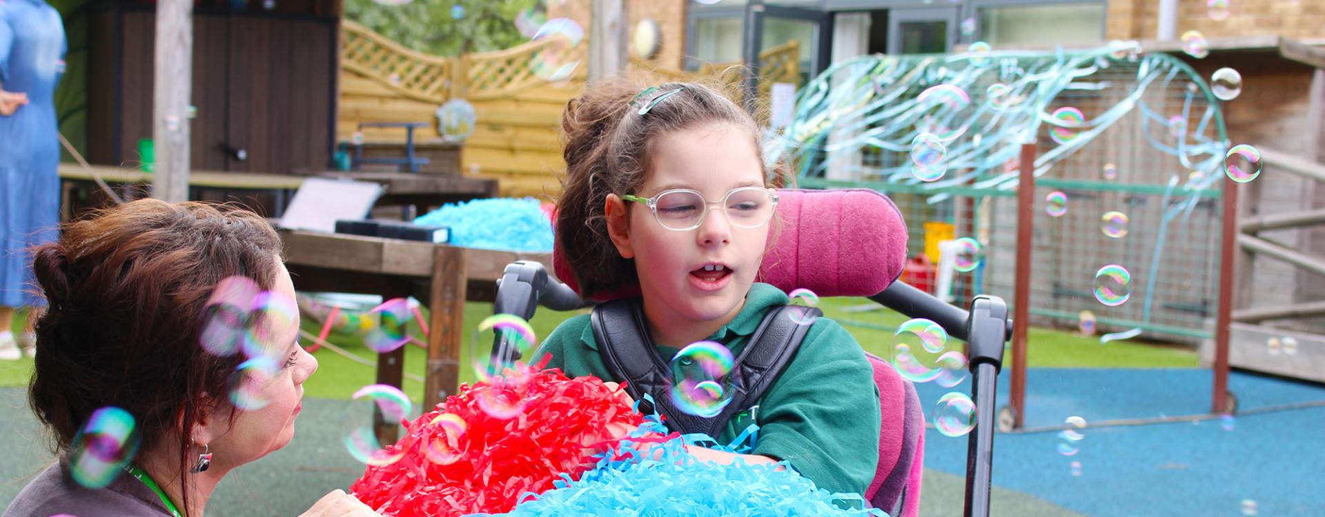 Treloar's primary student playing with colourful red and blue shredded paper while surrounded by floating soap bubbles outdoors.