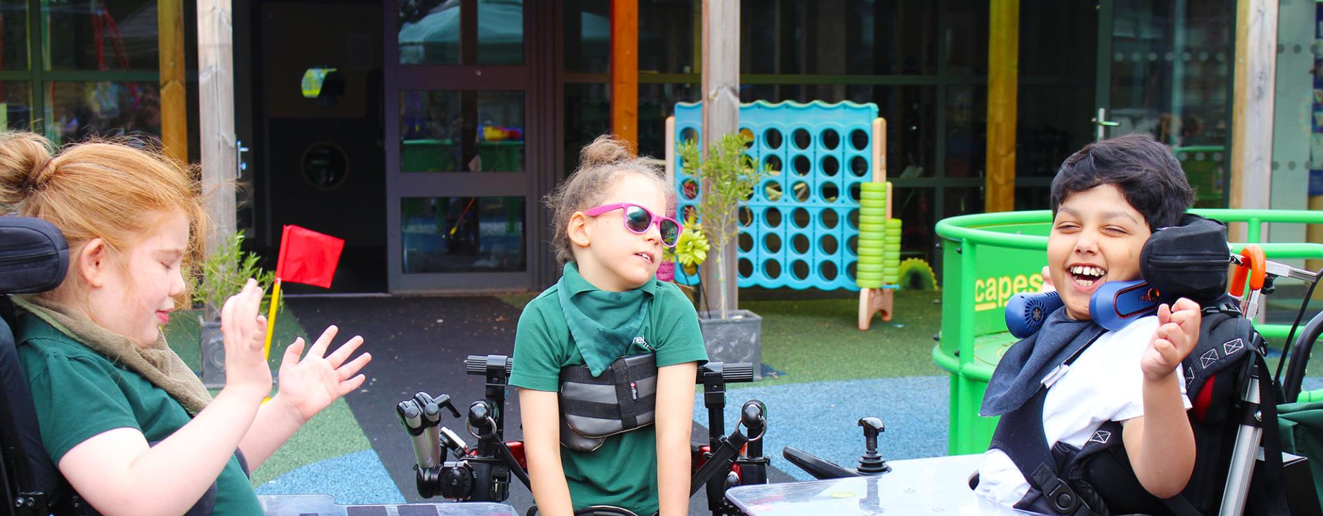 Three Treloar's primary students, two in wheelchairs, one in her standing frame, enjoy a sunny outdoor play area with games and laughter.