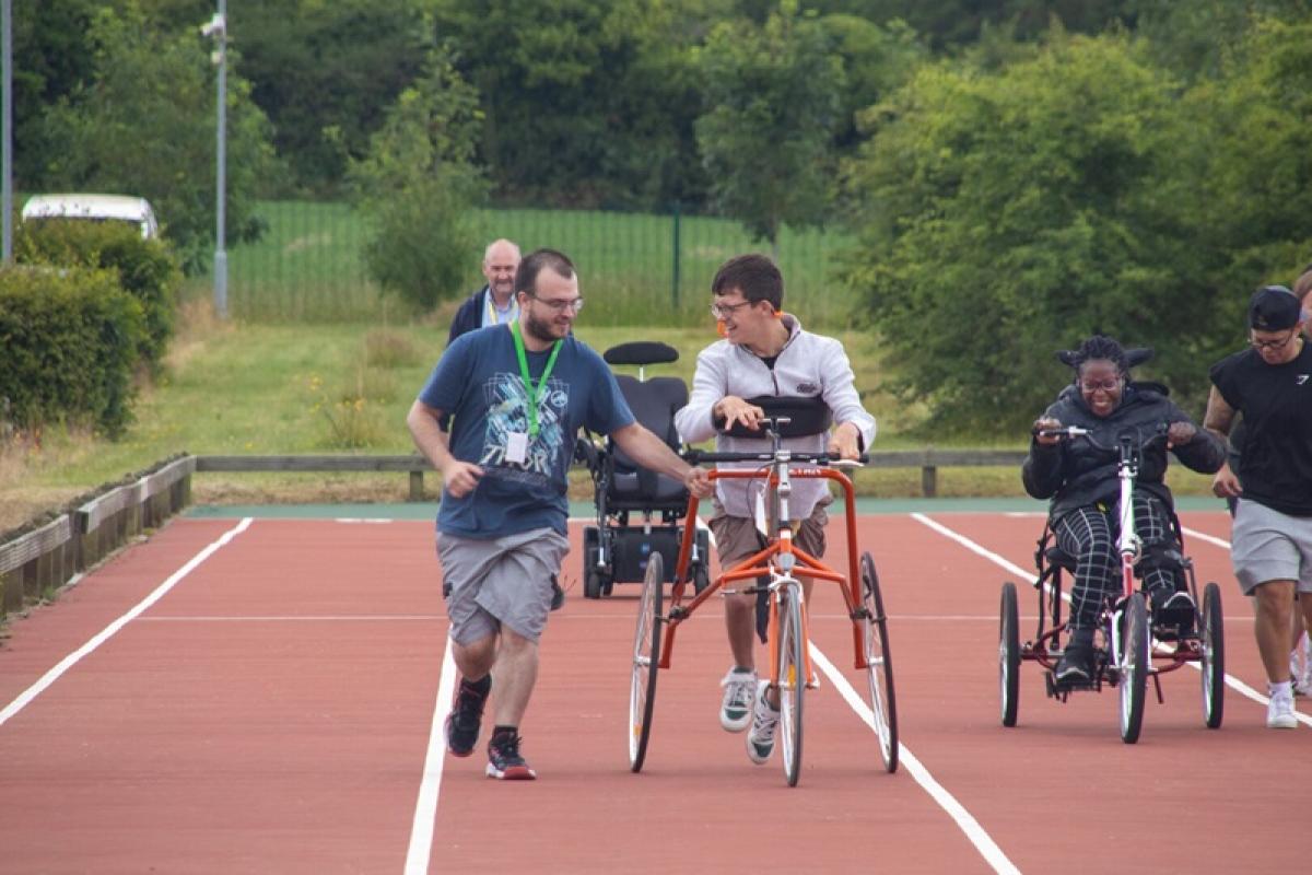 Treloar College student taking part in the annual Sports Day: he is using 3 wheeled walking frame supported by 2 members of staff.