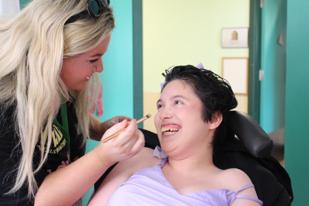 treloar's student support assistant applies makeup with a brush to Treloar College student wearing a lavender off-shoulder dress. They are laughing and having a good time.