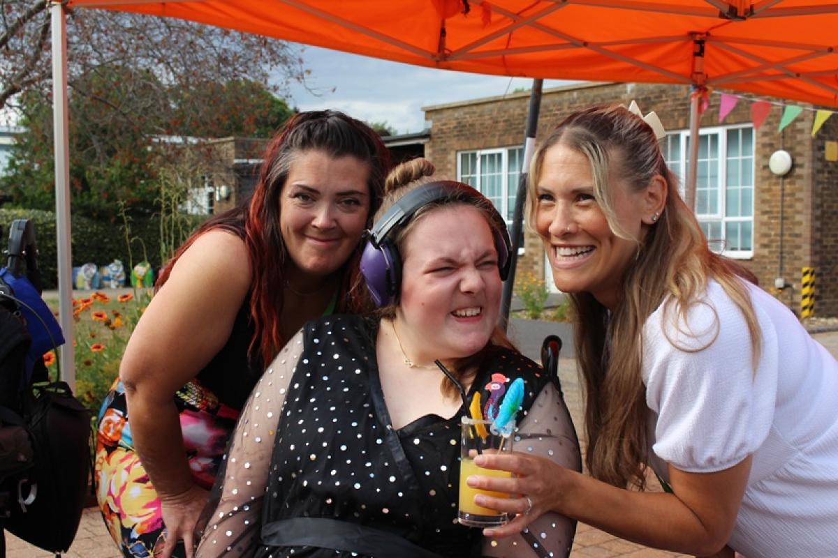 2 student support assistant with Treloar School student gathered under an orange canopy outdoors, during prom. One of them is holding a decorated glass of orange juice, all are smiling and enjoying the celebration.