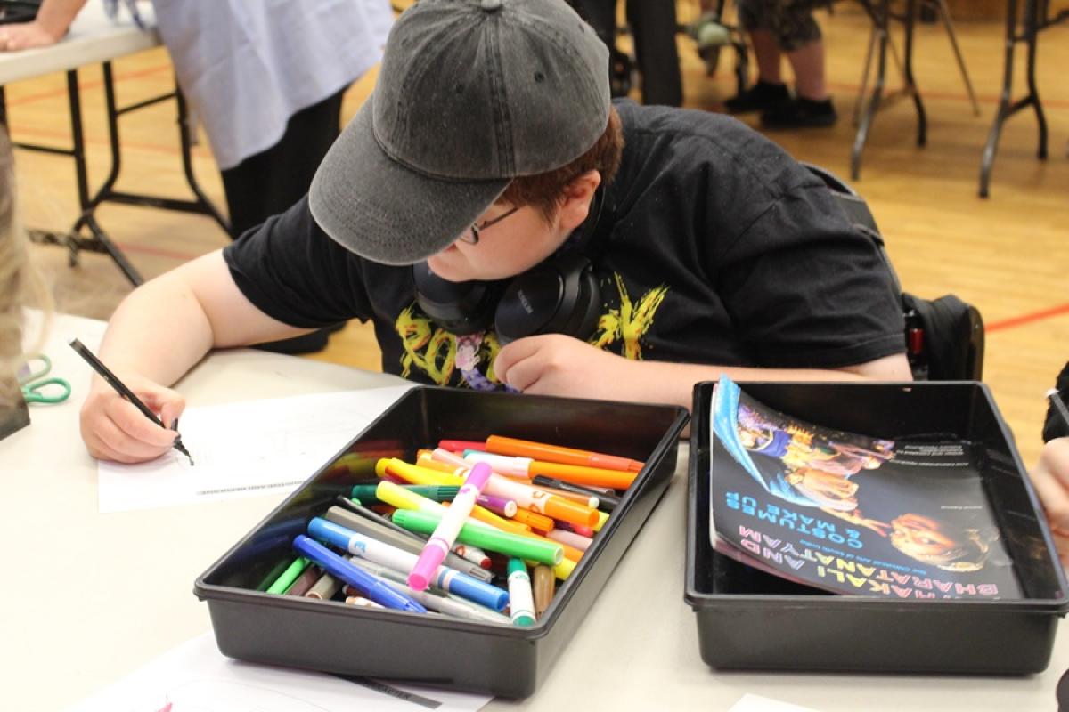 Treloar College student wearing a black cap, drawing with a black marker at a table filled with colorful markers and a book. 