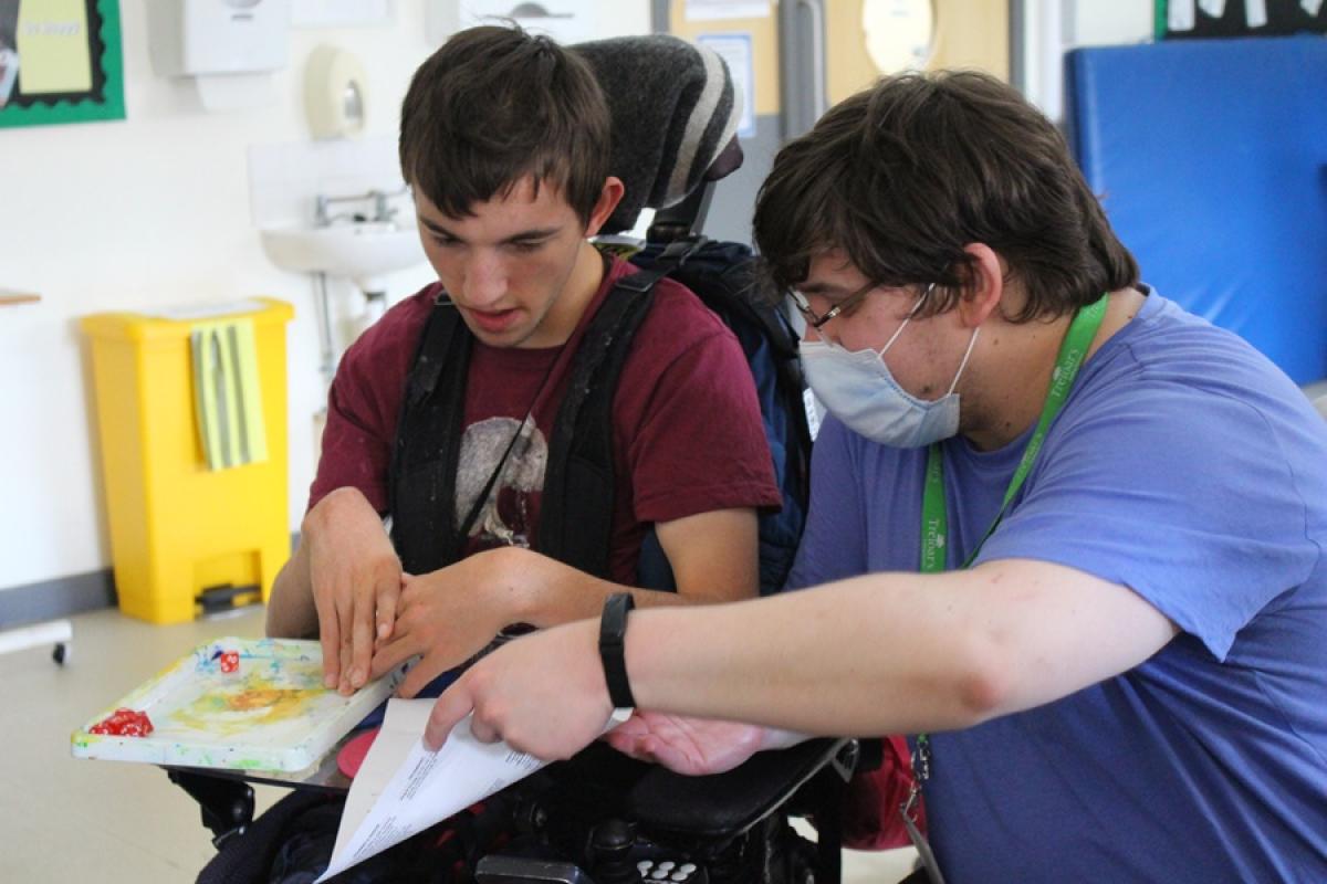 Treloar's student support assistant in a blue shirt is assisting Treloar College student with D&D board game by holding a paper with instructions and a colorful tray with a dice on.