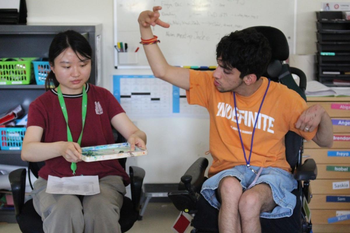 Treloar College student seated in a classroom with his assistant. The assistant is holding a board with a game dice on; the dice has just been dropped on the board by the student.