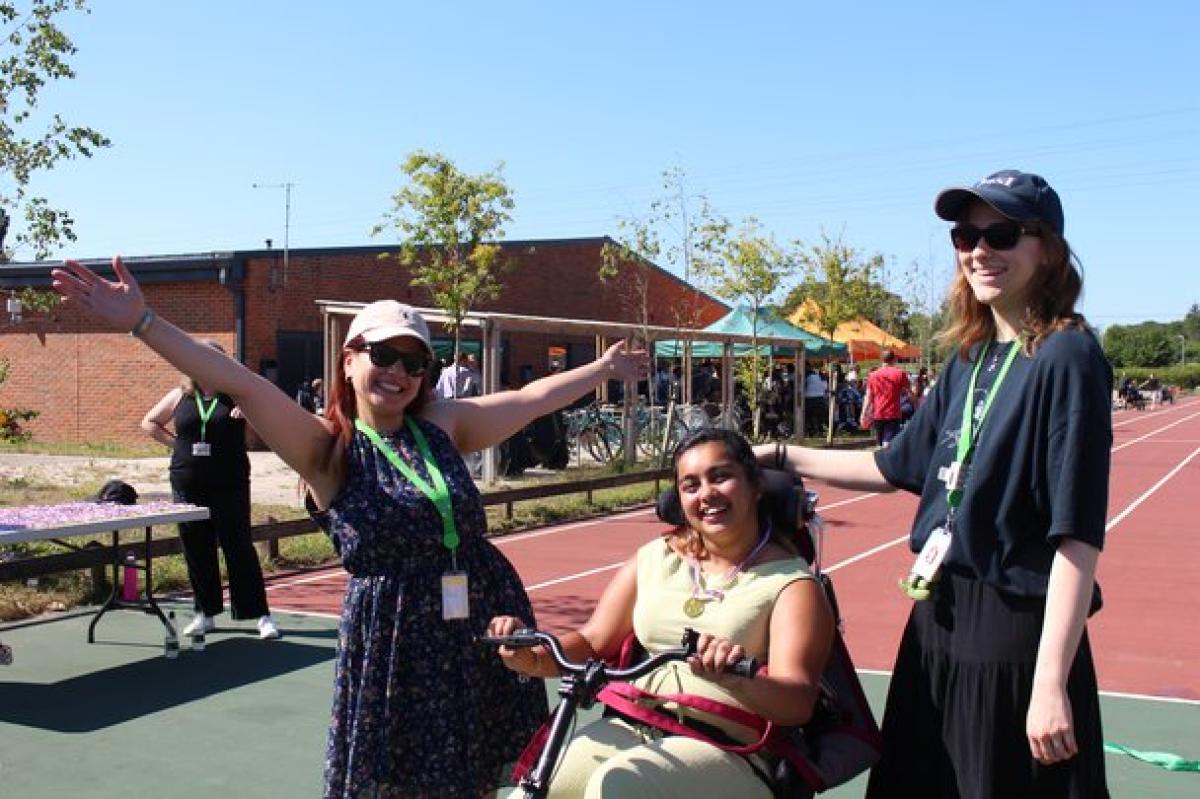 2 student support assistants and Treloar College student on a sunny day on a running track. The student in seated on a accessible trike and the assistants are standing nearby with arms outstretched and smiling.