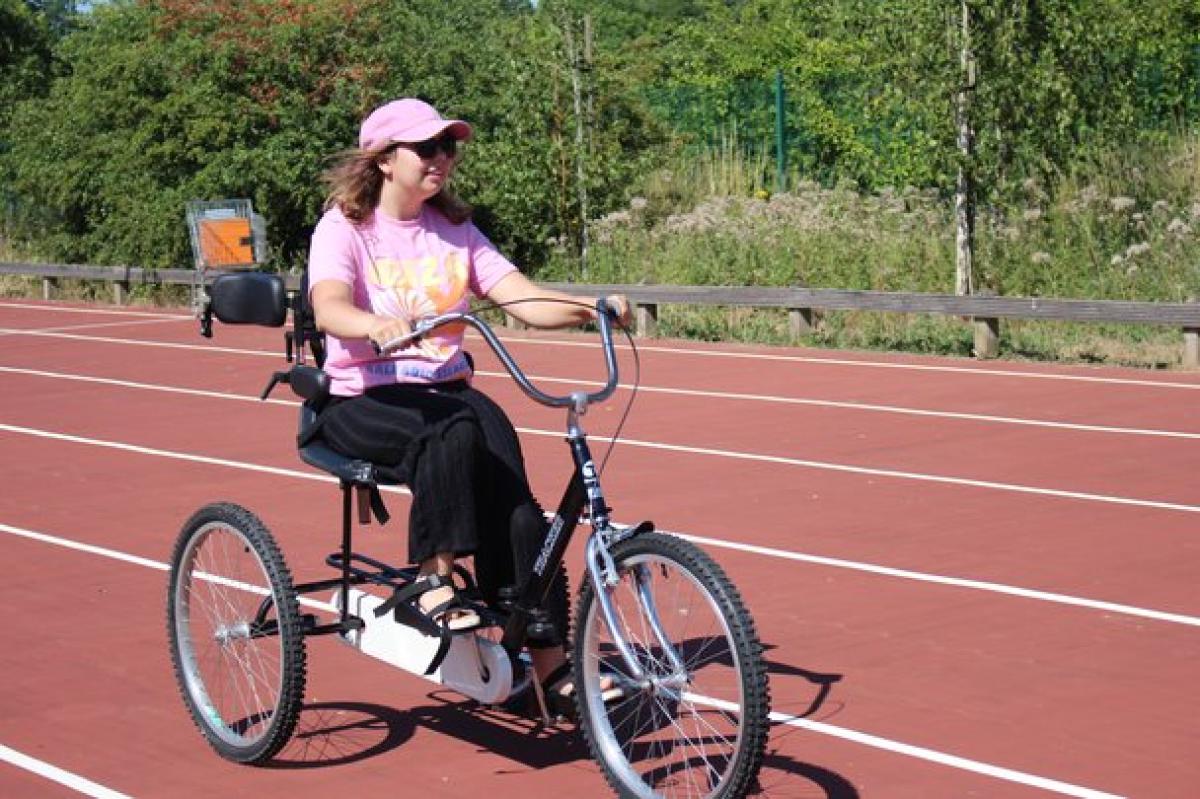 Treloar College student wearing a pink T-shirt and cap rides a black adult tricycle on a red outdoor track surrounded by greenery.