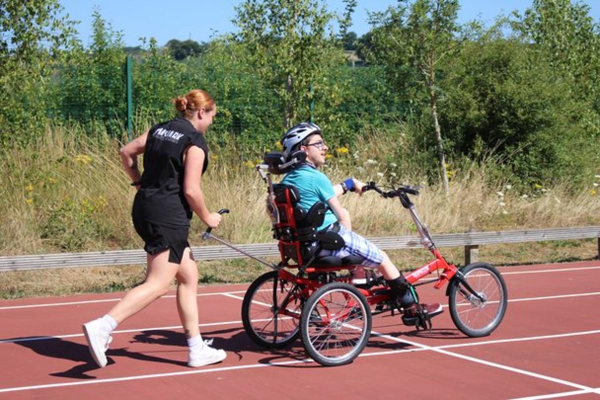 Treloar's student support assistant in athletic wear assists Treloar College student riding a red adaptive tricycle on an outdoor track surrounded by greenery.