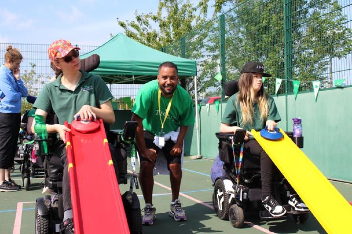 2 Treloar School students prepare to roll balls down colorful ramps during an outdoor Sports Day events on a sports court. Treloar's patron Martin Sinclair is posing for the photo, standing in between the 2 students.