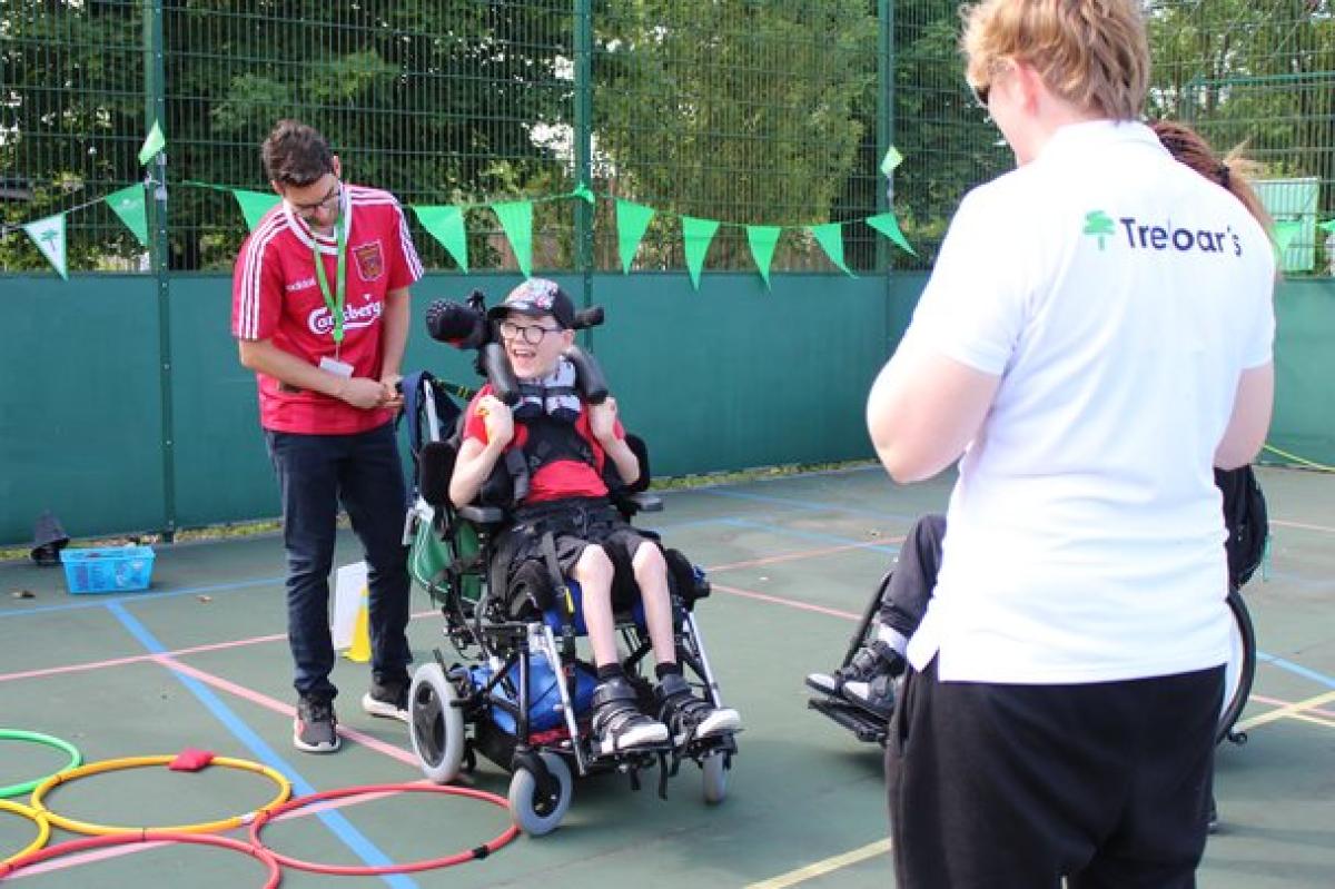Treloar School student participates in an outdoor activity on a sports court with colorful hoops on the ground; he is assisted by two student support assistants.