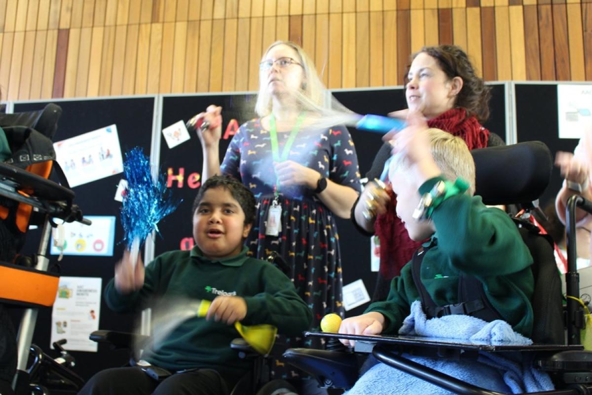 2 Treloar's students are holding and waving party accessories during a Makaton event; their assistants are behind them singing.