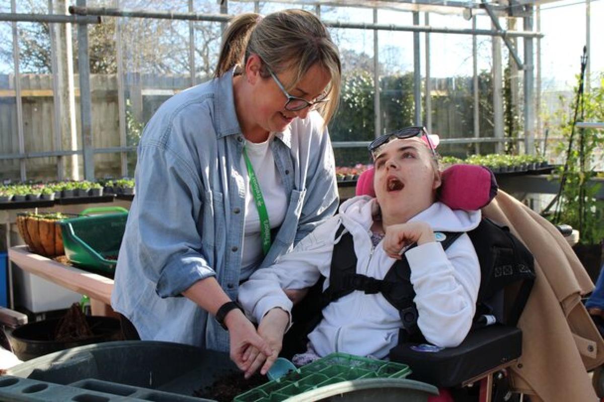 A support assistant assists Treloar's student with planting seeds in a greenhouse filled with gardening supplies.