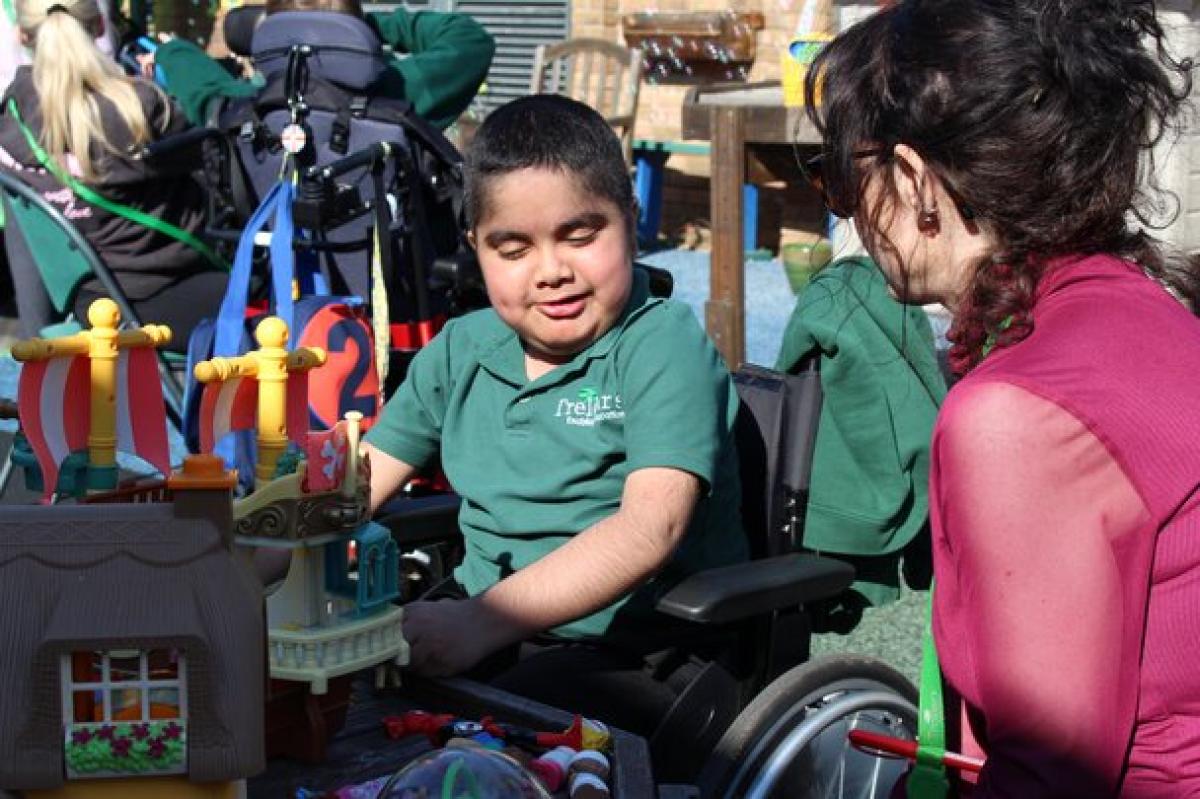 Treloar's primary student playing with colorful toys outdoors while his support assistant in a pink jacket watches nearby.