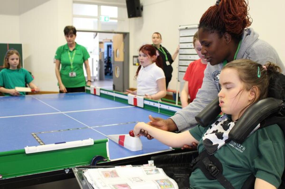 Treloar's student playing table cricket with assistance from her assistant. She is very focus on the ball as she prepares to roll it off the mini ramp. Other students are waiting.