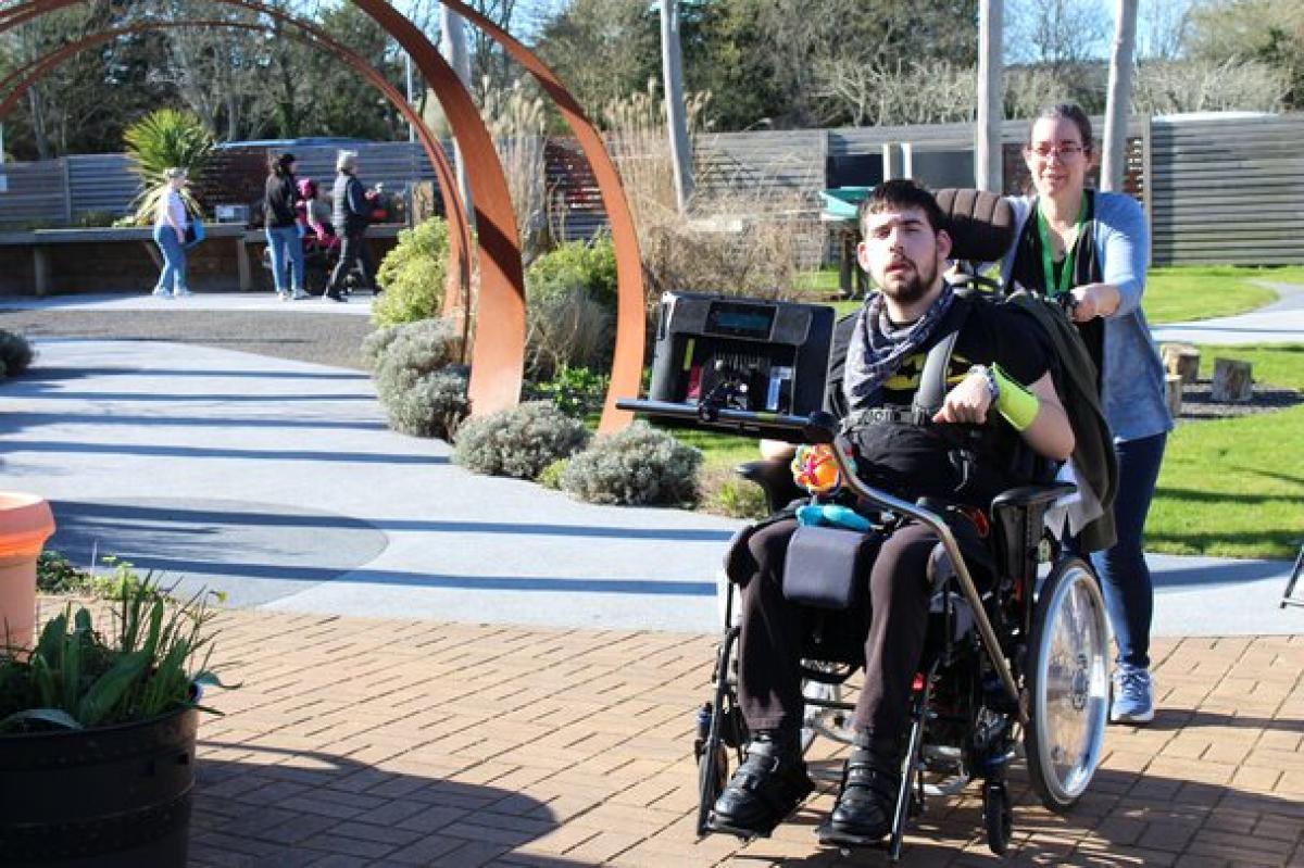 Treloar College student being pushed by his assistant along a sunny garden path with modern metal sculptures and greenery.