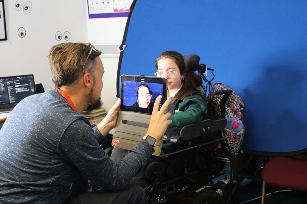 Man filming Treloar's student using a tablet in a classroom setting with a blue backdrop.