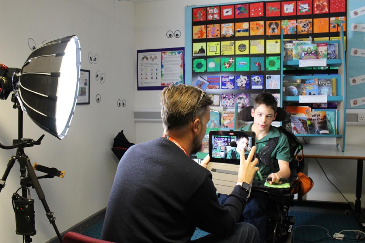 Man filming Treloar's student using a tablet in a colorful classroom with educational posters and a large studio light setup. 