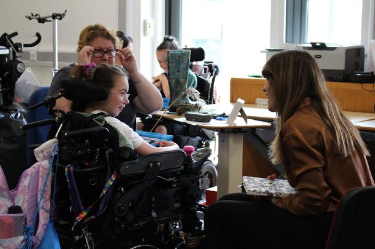 A visitor with long hair attentively interacts with Treloar's student inside a bright classroom with school equipment. The girl is smiling to the visitor and listening to her questions.