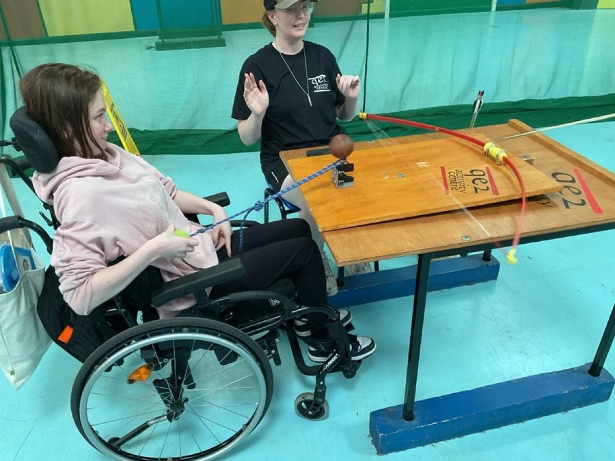 Treloar's students with a member of staff practising indoor archery. The student is using a rubber band that allows her to launch the arrow that is placed on a wooden table.
