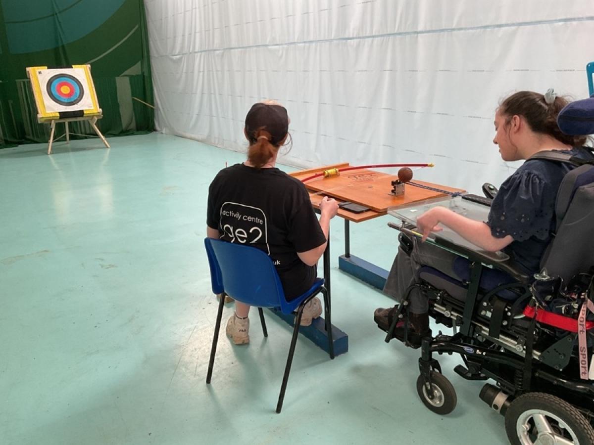 Treloar's students with a member of staff practising indoor archery. The student is aiming at a target downrange in a gymnasium.