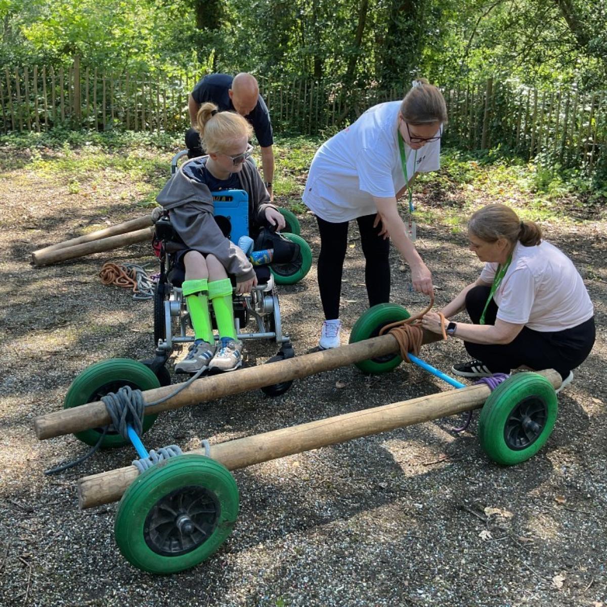 Treloar's student with bright green socks is assisted by three adults assembling a wooden go-kart outdoors on a gravel path. It's a sunny day and there are tall green trees in the background.