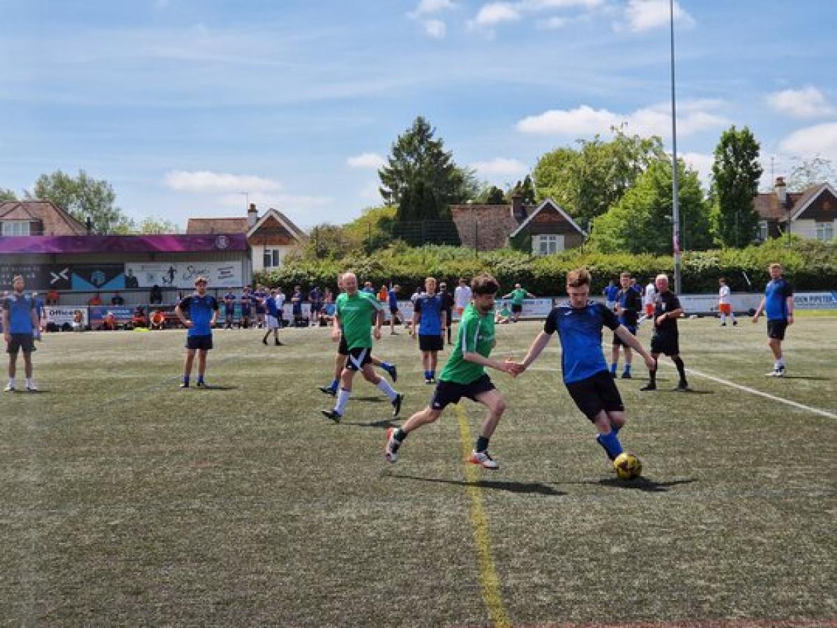 Football players compete on a sunny day at a local outdoor field with houses and trees in the background.
