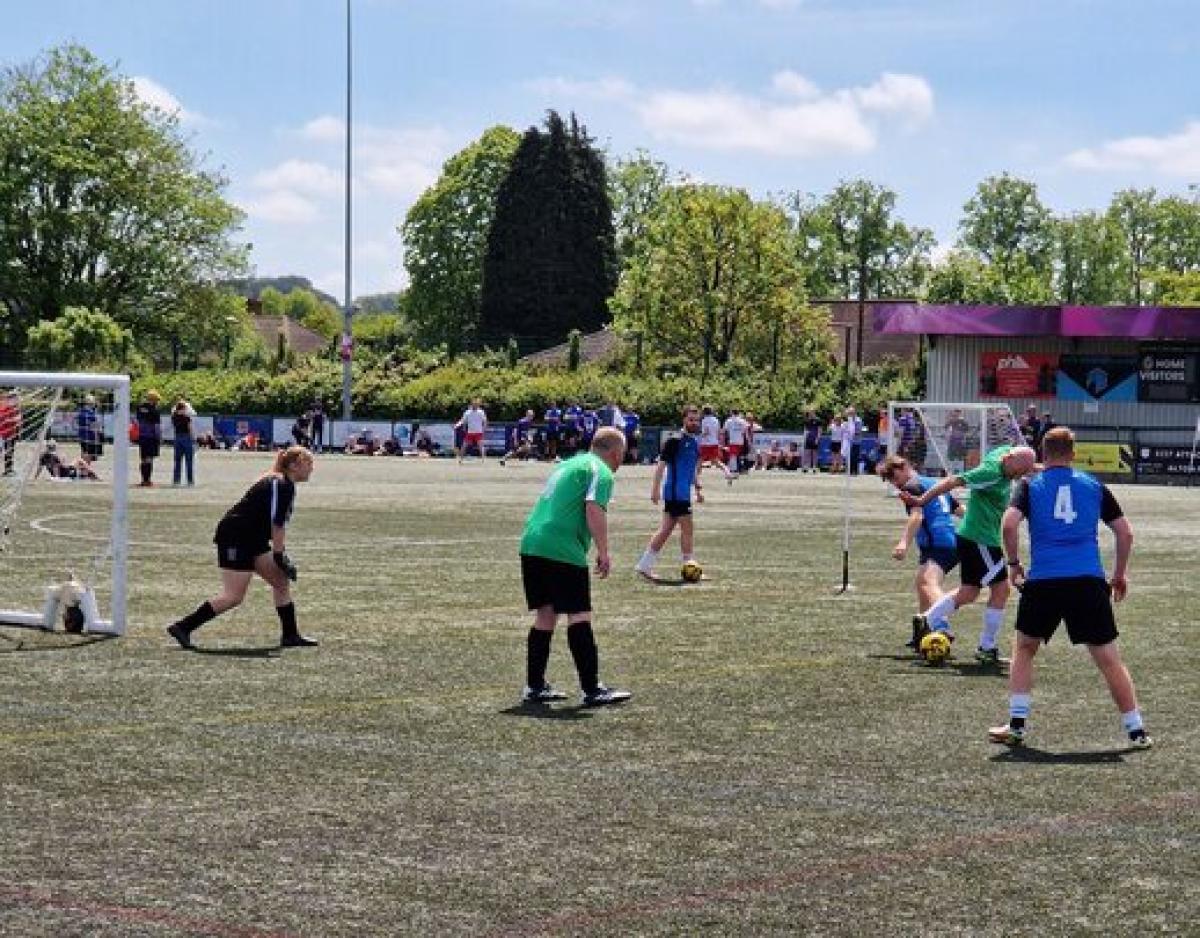 Amateur football match on a sunny day with players in green and blue jerseys competing near the goal.