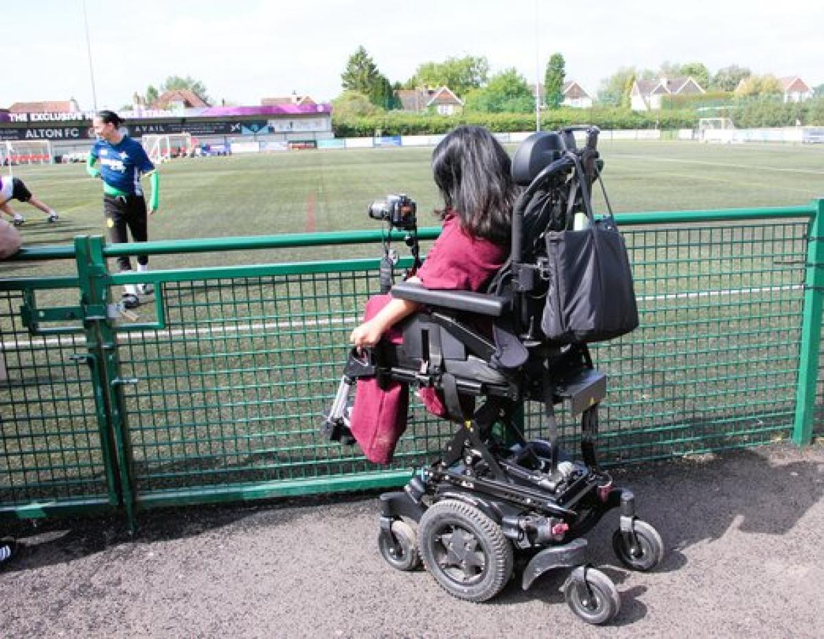 Treloar's student in her powered wheelchair that is raised, taking pictures of a football match from behind a green fence at a local sports field.