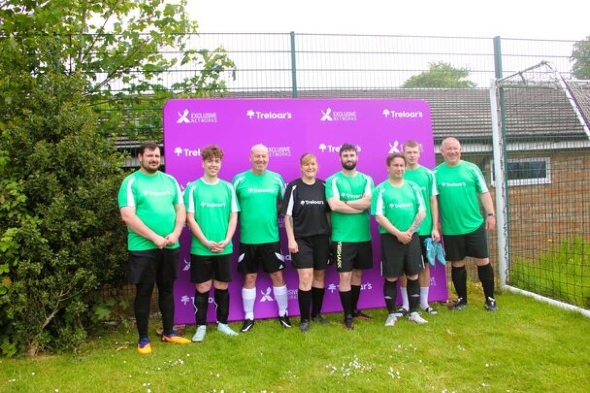 Seven football players from Treloar's in green and black uniforms stand on grass in front of a purple backdrop with sponsor logos at an outdoor field.