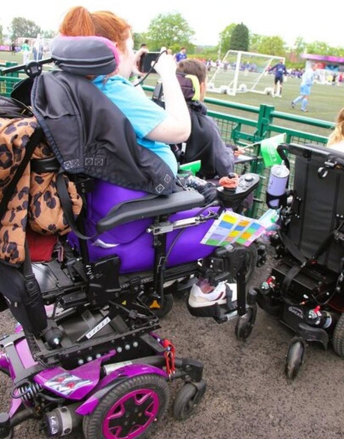 Treloar's student in her purple powered wheelchair that is raised to help taking photos at an outdoor football game with players visible in the background.