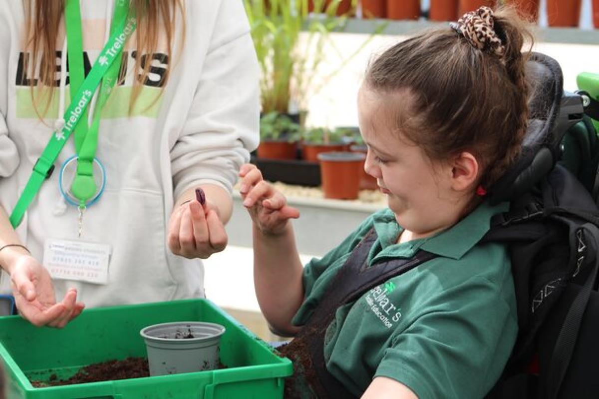 Treloar's student planting seeds in soil with assistance from her assistant. The student is wearing green uniform polo shirt and is very interested in the activity based in a greenhouse.