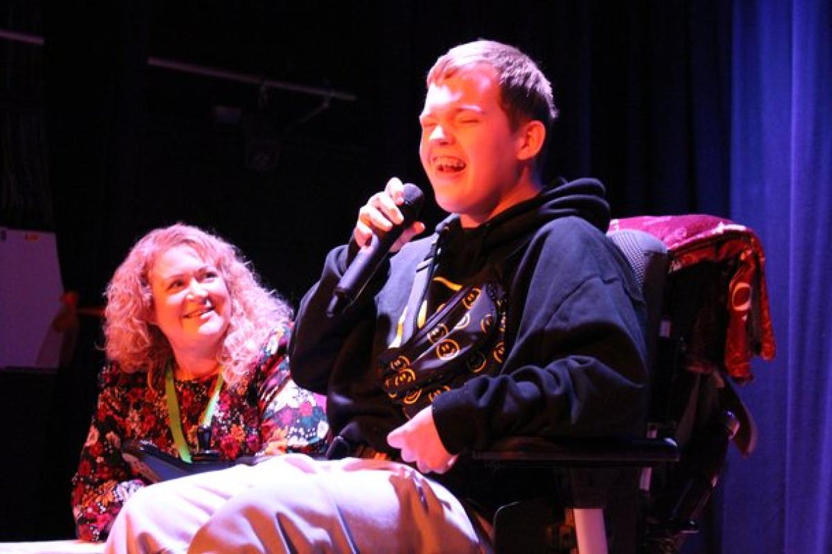 Treloar's student in a black hoodie singing into a microphone while on stage with his music teacher beside him. He looks like he is enjoying his performance. 