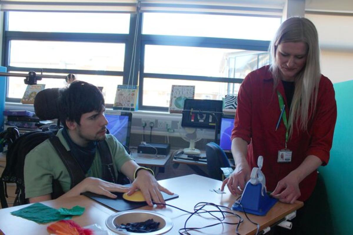 Treloar's student at a table using a switch to operate scissors while his assistant in a red shirt assists him with cutting up paper. They are in a bright room with large windows.