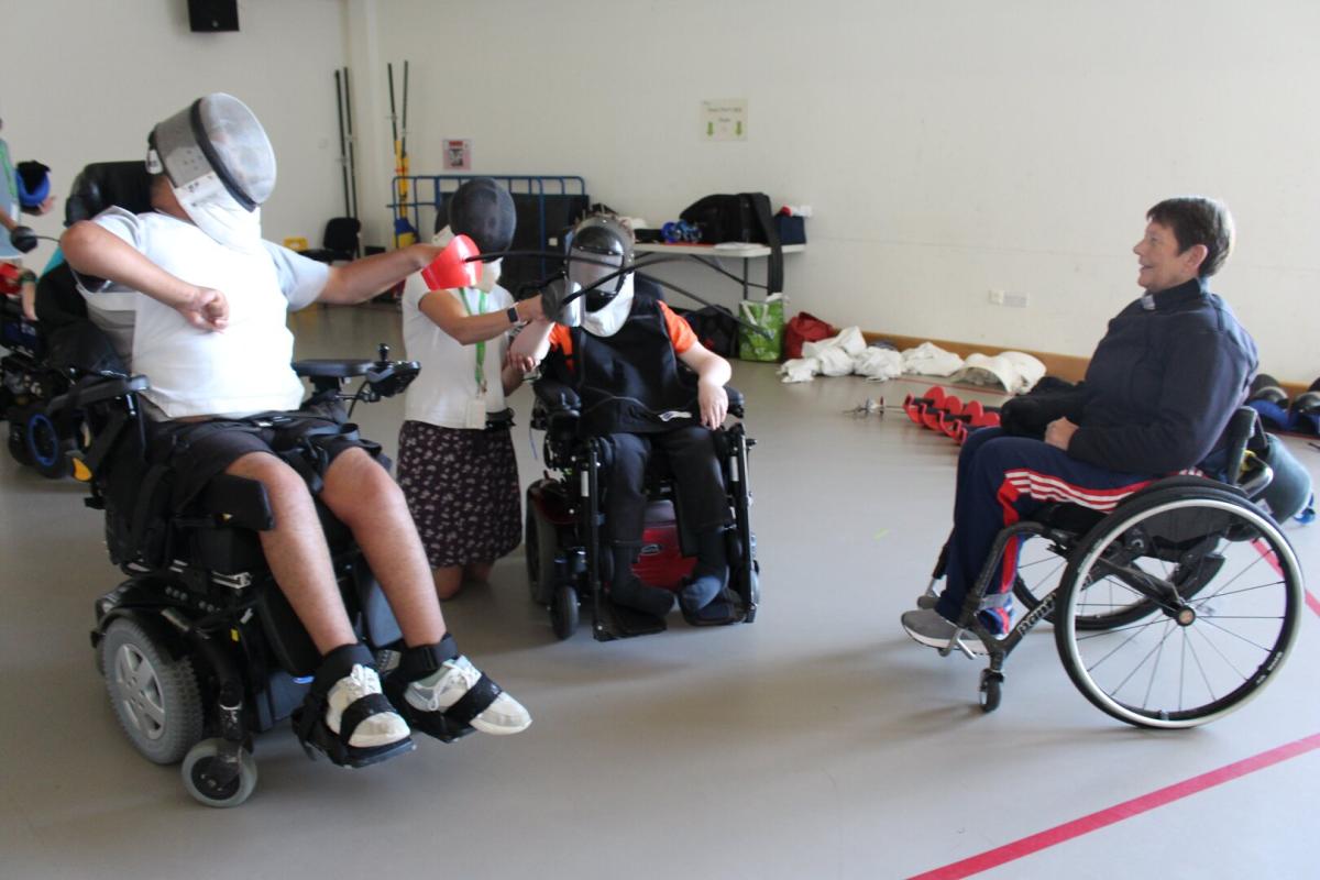 Students using wheelchairs try out fencing, wearing face masks and protective vests, observed by a coach