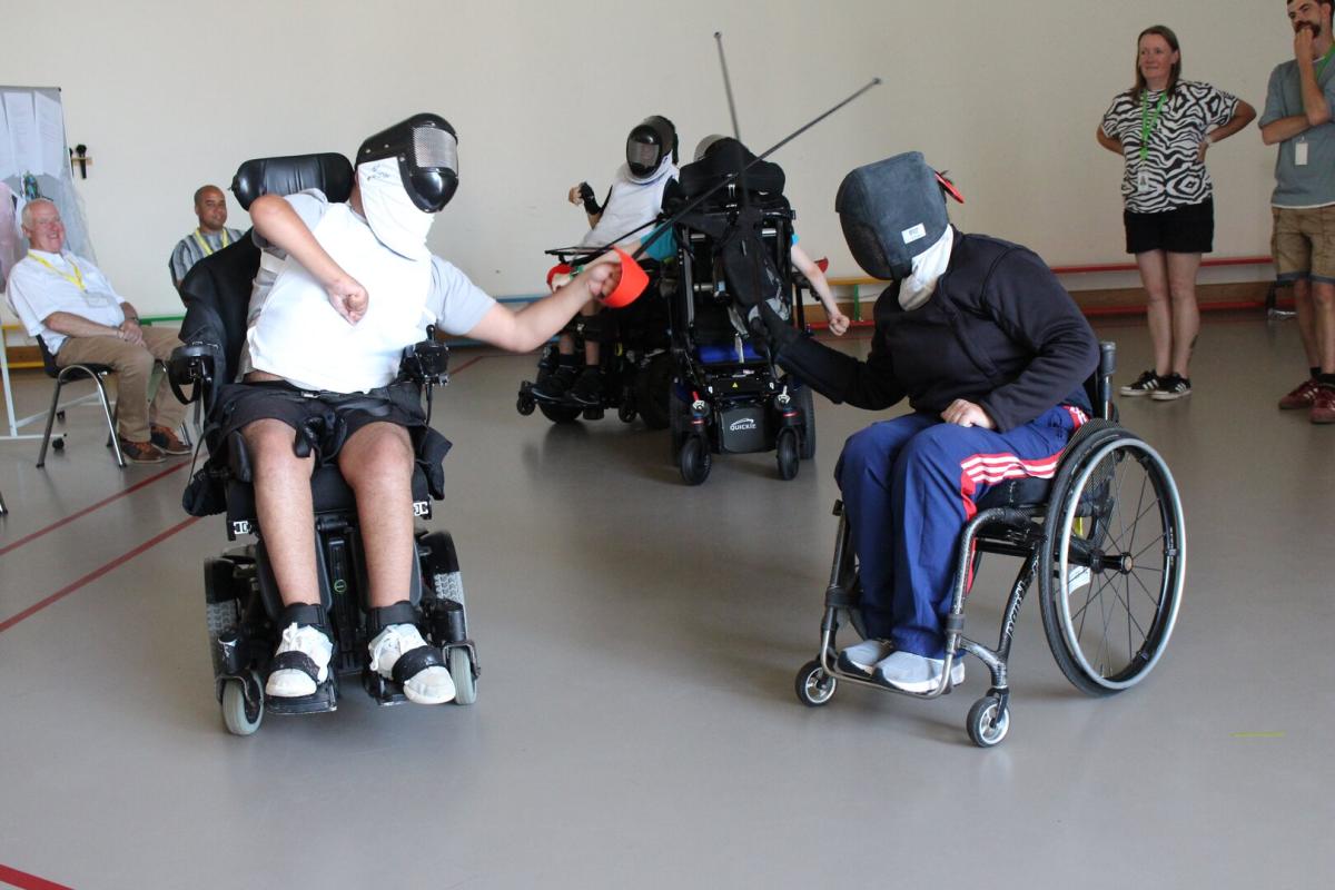 Two adults in wheelchairs try out wheelchair fencing, they are wearing face masks and protective vests, and their fencing weapons are locked in a cross shape.