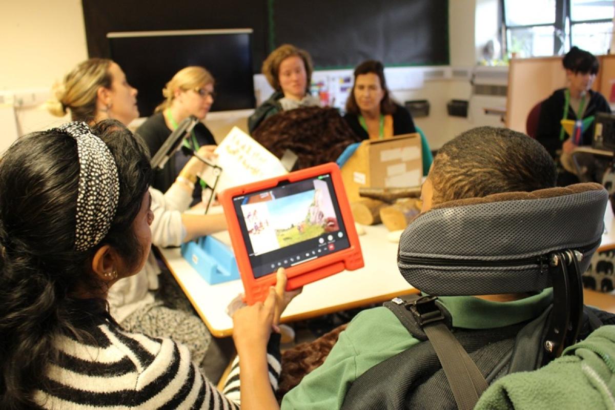 Treloar's students and staff gathered around a table, one person holding a tablet showing a colorful screen, engaged in discussion.