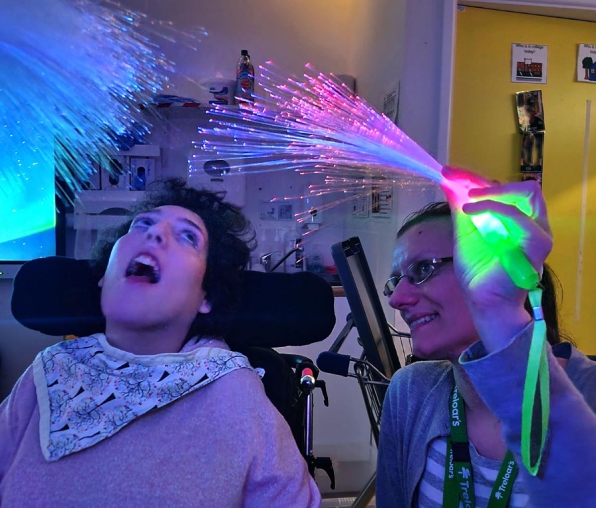 Treloar College student enjoying colourful fiber optic lights held by a smiling assistant in a sensory room. Lights are dimmed and the student is mesmerised by the fiber lights.