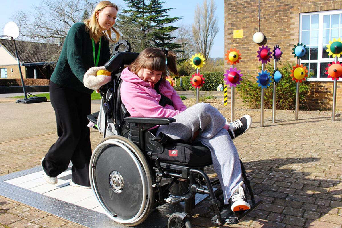 A student support assistant pushes a smiling Treloar College student outdoors near colourful flower-shaped musical instruments on a sunny day.