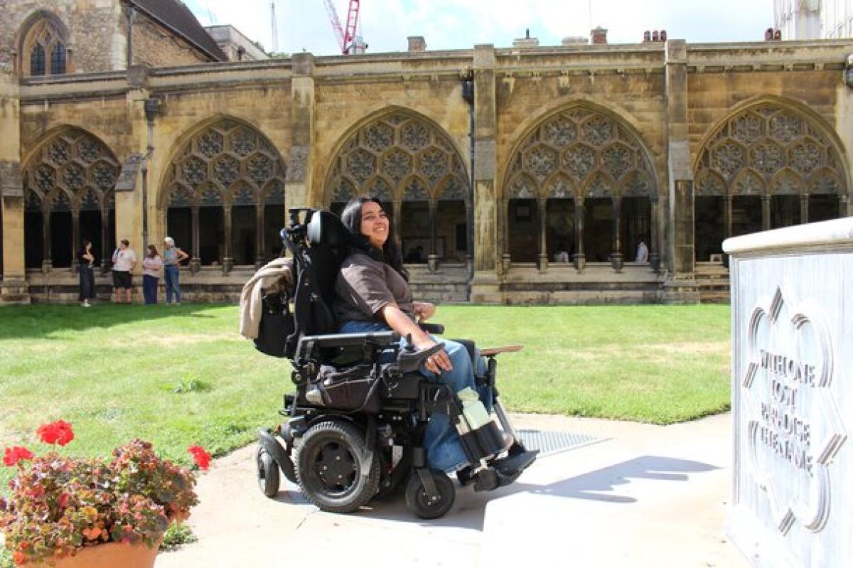 Treloar College student smiling outdoors near a historic stone building with arched windows and a grassy courtyard. It is a very sunny day.