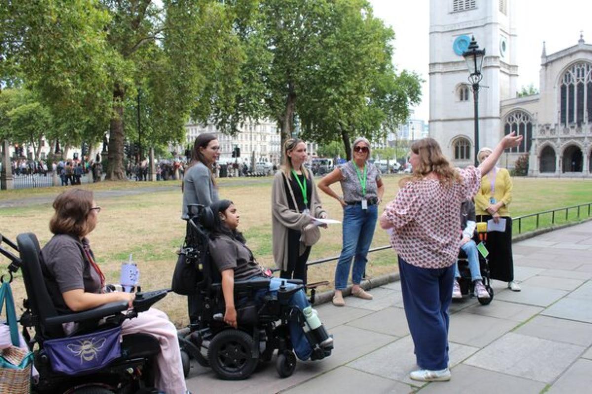 Group of people, including two in wheelchairs, attentively listening to a woman giving a tour outdoors near historic buildings and trees.