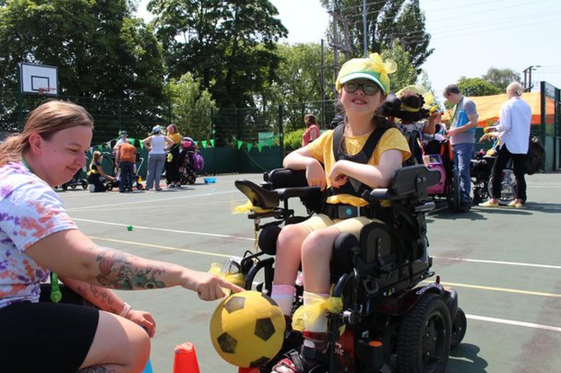 Treloar's primary student wearing yellow accessories plays with a yellow and black soccer ball on an outdoor court during a sunny day during Sports Day. Staff, students and families in the background.