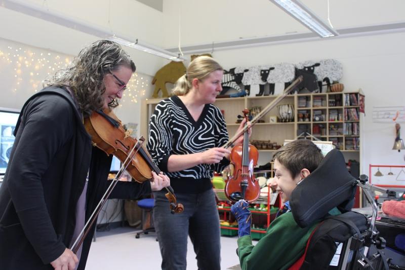 Kate Read, a professional viola player, is playing a viola, standing in front of Treloar's student who is smiling and enjoying the music; Treloar's Head of Music is standing next to them and is holding a violin.
