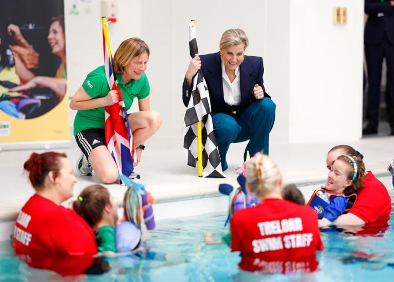 Her Royal Highness The Duchess of Edinburgh talking to Treloar's students who are in the pool with their assistants. The students have just finished a race and Her Royal Highness is holding a chequered flag.