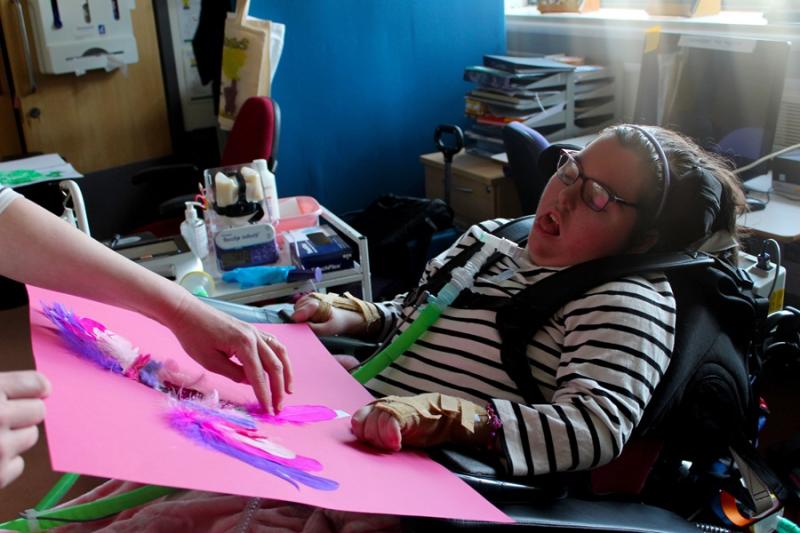  A student support assistant is glueing a pink feather onto a big pink paper after being directed to do so by a Treloar's student who is looking at the stage prop.
